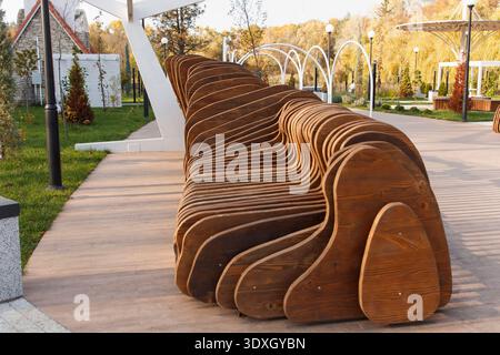Una moderna panca scultorea realizzata in legno marrone a strati si trova su un passaggio pedonale in un parco paesaggistico. Le linee principali della progettazione parametrica creano una dinam Foto Stock