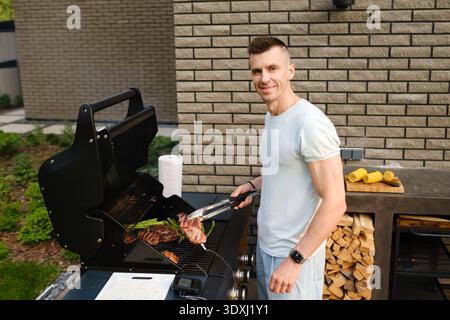 Un uomo cucina diversi tipi di carne su una griglia in un cortile. Sta accanto alla griglia e tiene le pinze sorridenti. E' una giornata di sole con il verde Foto Stock
