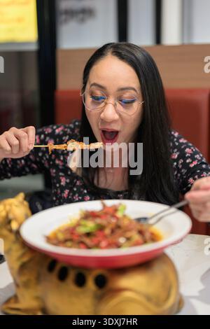 Felice donna asiatica che indossa gli occhiali che mangia un colorato piatto asiatico cinese speziato nel ristorante Foto Stock