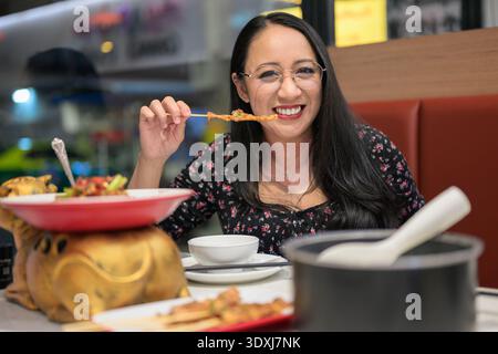 Donna felice che tiene in mano lo spiedino satay mentre cena all'interno Foto Stock