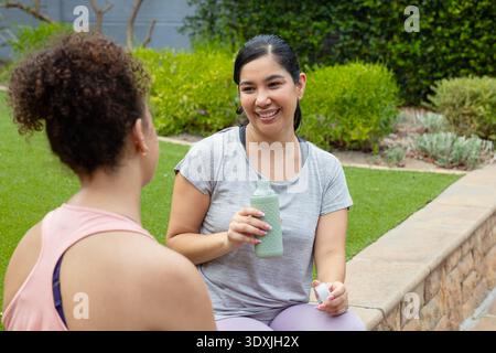 Diverse amiche in abbigliamento sportivo sedute su un muro di pietra che regge una bottiglia verde e un berretto, chiacchierando Foto Stock