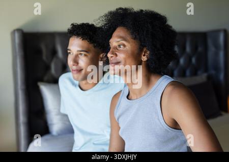 Due uomini afroamericani seduti sul letto con testiera in camera da letto, che indossano una canotta grigia Foto Stock
