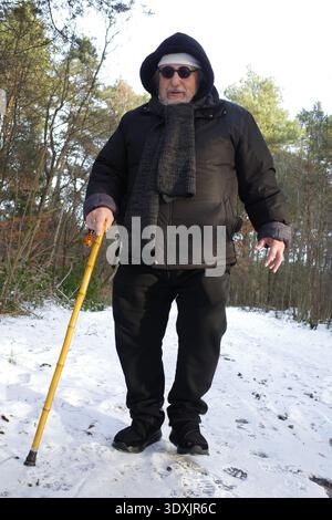 Un anziano cammina su un sentiero innevato in una zona boscosa. Indossa un cappotto invernale nero, cappuccio, sciarpa e occhiali da sole. Ha una CA gialla che cammina Foto Stock