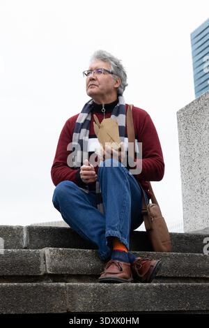 Uomo anziano seduto su gradini di pietra che regge scatola da asporto e tovagliolo, sciarpa, occhiali, borsa abbronzante Foto Stock