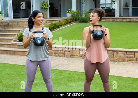 Diverse amiche che indossano kettlebell da 5 kg in abbigliamento sportivo e smartwatch in giardino Foto Stock