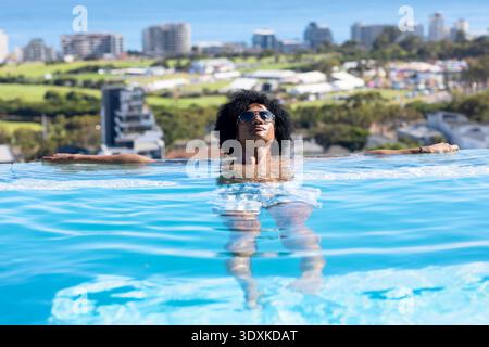 Uomo africano adulto che si rilassa nella piscina a sfioro sul tetto, indossa occhiali da sole e baule leggere Foto Stock