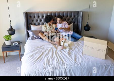 Papà afroamericani che reggono il telefono, puntano e si siedono su un letto con i neonati, scatola donazioni Foto Stock