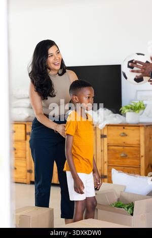 Mamma ispanica che tiene la spalla di un ragazzo afroamericano, sorride, mano che offre palla da calcio vicino ai box Foto Stock
