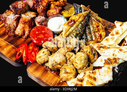 Vari tipi di carne alla griglia sono disposti su un piatto di legno. I contorni includono peperoni rossi, pane e salse. Foto Stock