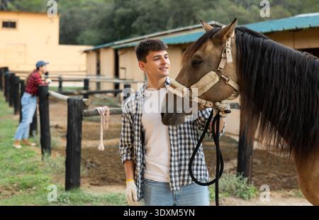 Giovane ragazzo positivo allevatore in camicia a quadri e guanti che preparano l'equitazione nel paddock a stalla Foto Stock