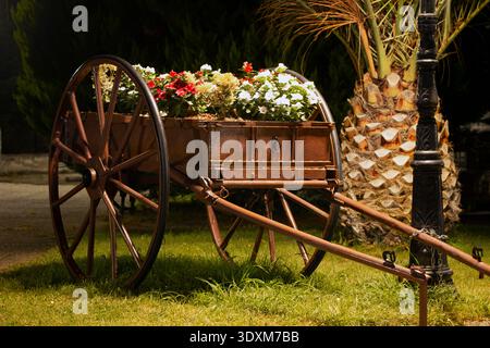 Carrello in metallo rustico e legno usato come fioriera in giardino con fiori colorati palme e calda illuminazione notturna decorativa all'aperto. Foto Stock