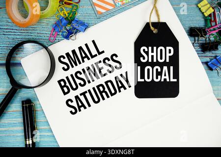 Small Business Saturday testo su carta per appunti e Shop Local con cartellino di prezzo circondato da forniture per ufficio e lente d'ingrandimento su un surf in legno blu Foto Stock