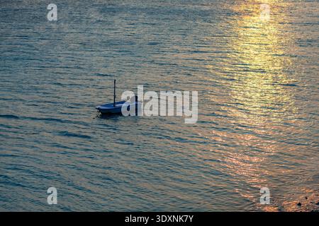 Il tramonto risplende sulle acque calme. La piccola imbarcazione si sposta dolcemente sul mare ondulato. La figura sagomata è seduta, ferma e silenziosa. La luce dorata balla attraverso la S. Foto Stock