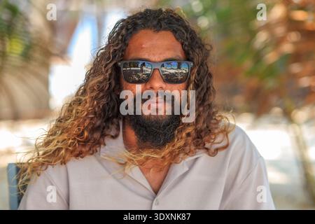 L'uomo con lunghi capelli ricci e barba indossa occhiali da sole. Siede all'aperto, rilassato e calmo. La luce filtra attraverso gli alberi dietro di lui. Ti consigliamo un abbigliamento informale Foto Stock