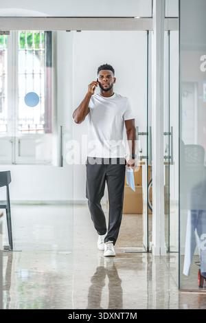 Un giovane vestito con una t-shirt bianca e pantaloni neri cammina con sicurezza attraverso un corridoio dell'ufficio, parlando sul suo smartphone e tenendo i documenti che ho Foto Stock
