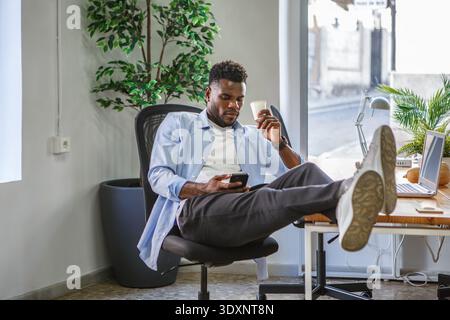 Un giovane uomo siede comodamente su un'elegante sedia da ufficio con un caffè in mano e consulta il suo smartphone in modo noncurante. Foto Stock