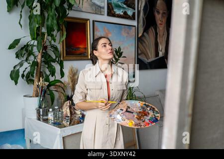 Artista donna positiva che guarda la tela con un'espressione ispirata, godendo della terapia artistica. Foto Stock
