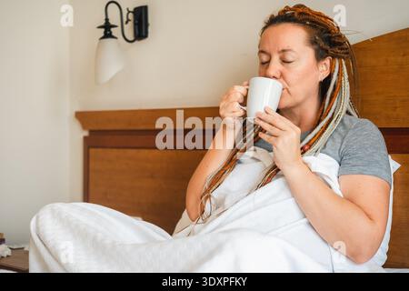 Donna di mezza età che beve caffè mattutino a letto. La donna con i dreadlocks sulla testa sorride contentosamente. || modello rilasciato Foto Stock