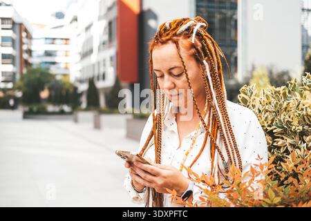 Una donna moderna di mezza età con dreadlock guarda lo schermo dello smartphone. La donna usa il telefono per strada. Donna moderna in città || modello rilasciato Foto Stock