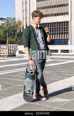 Uomo sui 20 anni in piedi sul ponte sul tetto che tiene lo skateboard con le cuffie e controlla lo smartphone. Urbano, parcheggio, tabellone, zaino, auricolari, telefono, luce solare || modello rilasciato Foto Stock