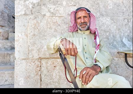 Un uomo anziano è seduto contro un muro di pietra ad Aleppo. Indossa abiti tradizionali e regge un bastone da passeggio. Foto Stock