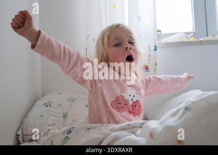 Adorabile bambina di 4 anni che sbadiglia e si allunga dopo essersi svegliata in un'accogliente camera da letto illuminata dal sole Foto Stock