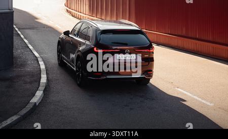 Mazda CX-90 rosso scuro su una strada industriale illuminata dal sole. Un'immagine retrovisiva ad alto angolo di un SUV Mazda CX-90 rosso scuro che guida lungo una parete ondulata rossa curva Foto Stock