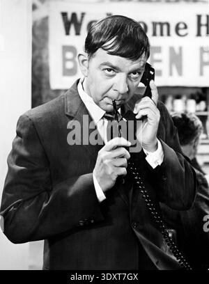 Questa fotografia in bianco e nero del 1970 cattura l'attore Woodrow Parfrey come il suo personaggio Braden Pyle, dalla serie televisiva "Mayberry R.F.D.". Viene mostrato tenendo un ricevitore telefonico all'orecchio mentre tiene in bocca una penna o un piccolo oggetto, con uno striscione visibile sullo sfondo. Foto Stock