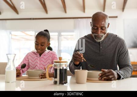 Uomo maturo afroamericano e ragazza giovanile che mangiano al bancone di casa con stampa francese, felpa con cappuccio rosa Foto Stock