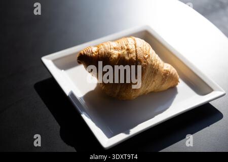 Croissant flaky dorato servito su un piatto quadrato bianco al moderno tavolo da bar. Foto Stock