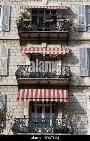 Classico balcone di Nizza con tende a righe rosse e bianche, ringhiere in ferro battuto nero ornato e piante in vaso su una storica facciata in pietra. Nizza, Francia Foto Stock