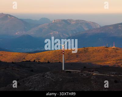 La vista aerea di una sola turbina eolica si erge in alto sullo sfondo di colline ondulate e montagne lontane, baciate dal caldo bagliore del sole che tramonta Foto Stock