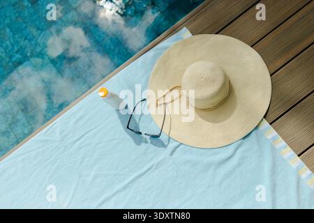 Cappello da sole in paglia intrecciata, occhiali da sole neri e crema solare si appoggiano su un asciugamano azzurro a bordo piscina Foto Stock