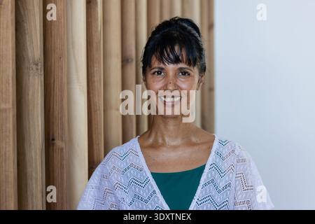 Donna matura che sorride in corridoio con spranghe di legno, parete pallida, top color ottanio e scialle bianco Foto Stock