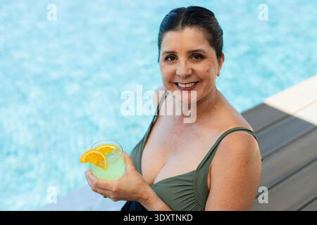 Donna matura in costume da bagno verde seduta sulla terrazza della piscina sorridente, con cocktail con zeppe arancioni Foto Stock