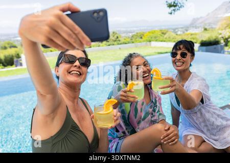Diverse amiche femminili sedute in piscina a fare selfie con il telefono che sorregge cocktail con occhiali da sole Foto Stock