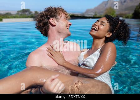 Coppia che abbraccia e ride mentre galleggia nella piscina a sfioro con costume da bagno a righe e occhiali da sole Foto Stock