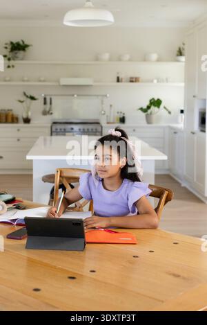 Preadolescente afroamericana seduta al tavolo della cucina usando un tablet con stilo e note di scrittura Foto Stock
