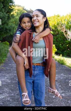 Madre afroamericana che porta la figlia giovane sul retro, camminando lungo il sentiero pavimentato del giardino in jeans Foto Stock
