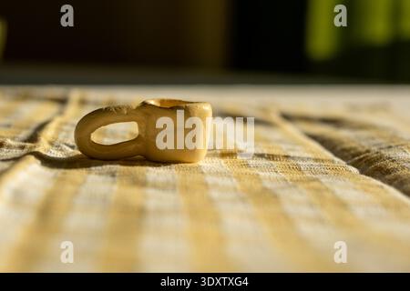 Tazza di argilla in miniatura su tessuto a scacchi giallo alla luce calda del sole. Foto Stock
