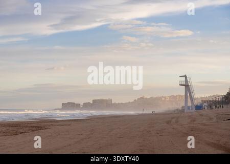 Una tranquilla spiaggia di Torrevieja, in Spagna, caratterizzata da una torre bagnino sulla sabbia, con onde che si infrangono dolcemente e un paesaggio urbano lontano sotto un morbido sk Foto Stock