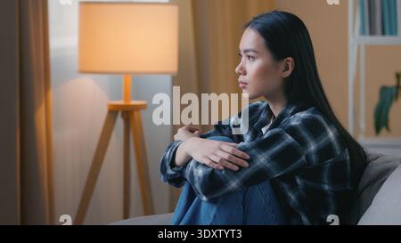 Vista laterale donna asiatica triste seduto sul divano a casa incerto stress mentale infelice ragazza coreana turbata depressa rammarico errore psicologico Foto Stock