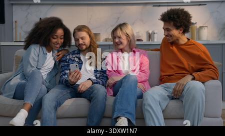Uomo caucasico a casa che mostra divertente video amici di cellulare seduti sul divano ridendo insieme gruppo multietnico uomini donne ragazze africane Foto Stock