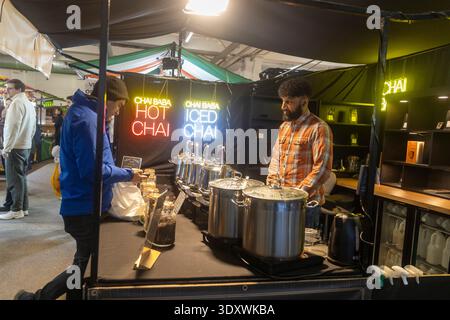 Londra, Inghilterra, 12 dicembre 2025, Un venditore in un mercato di Street food che serve tè caldo in una stalla chiamata Chai Baba con insegne al neon. Foto Stock