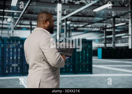 Scienziato informatico che cammina nel data center eseguendo linee di codice sui notebook, risolvendo i problemi dei montaggi su rack. Sviluppatore di software afroamericano che utilizza script di programmazione su un dispositivo notebook nella sala server Foto Stock