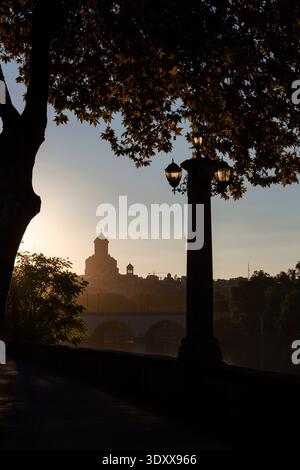 Vista all'alba della sagoma della Cattedrale della Santissima Trinità con lampione stradale vintage e ponte sul fiume kurá a Tbilisi, Georgia Foto Stock