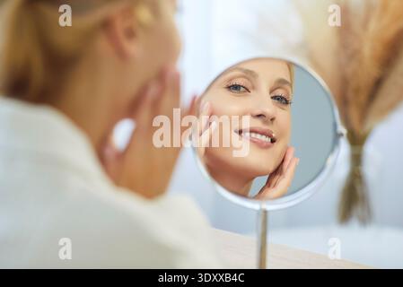 Donna che guarda allo specchio, tocca la pelle del viso con un sorriso contento Foto Stock
