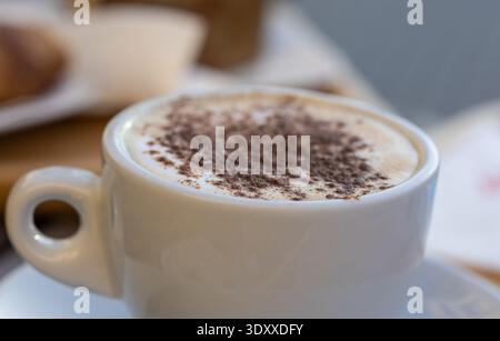 primo piano di un cappuccino cremoso in una tazza di ceramica bianca con spolveratura di cacao in polvere, sfondo sfocato per la colazione con croissant e un'estetica accogliente al mattino Foto Stock