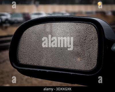 Primo piano di gocce di pioggia sullo specchio laterale dell'auto con sfondo sfocato in una giornata di pioggia Foto Stock