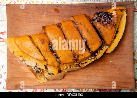 Vista ravvicinata del Martabak Manis a fette su un piatto di legno, che mostra una consistenza spessa e una ricopertura di cioccolato fuso su uno sfondo a motivi geometrici. Foto Stock